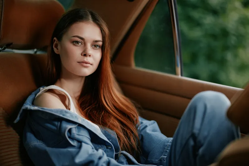 Young woman with long hair in denim attire sitting in a stylish car, capturing a moment of serenity and contemplation in a lush green environment.