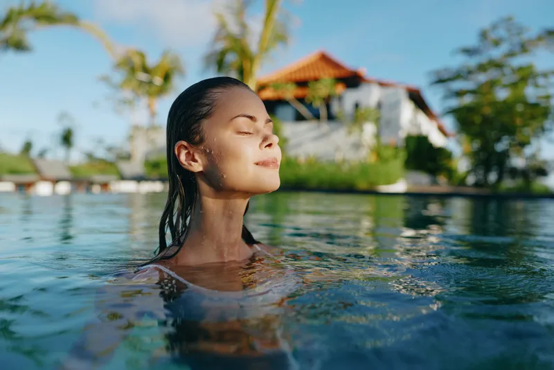 Relaxed woman emerging from a sunlit pool, eyes closed, serene smile, tropical resort backdrop, clear water, gentle breeze, natural colors, vacation mood and peaceful mood