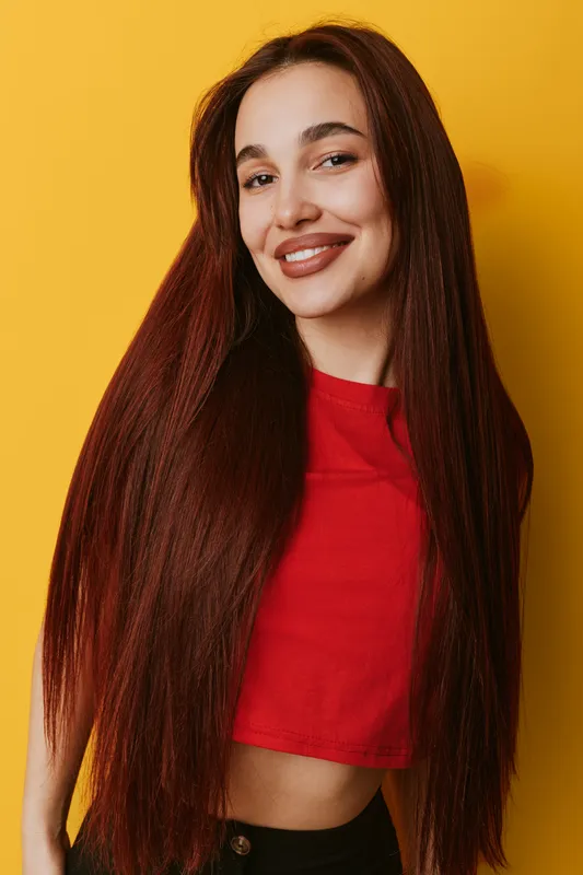 Young woman with long straight hair wearing red crop top smiling and posing against yellow background. Portrait of happy female with natural makeup and casual style indoors.