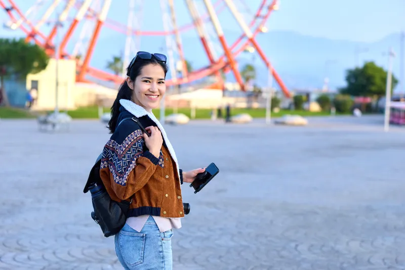 Ferris wheel backdrop at a sunny amusement park, smiling traveler with backpack, casual jacket and denim, exploring outdoors with a device in hand, vivid lifestyle moment for travel and fashion