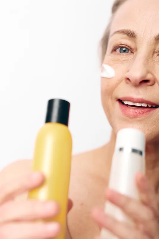 Mature woman with natural skin applies white cream on face holding yellow and white cosmetic bottles. Concept of skincare, anti aging, beauty routine, and facial hydration care.