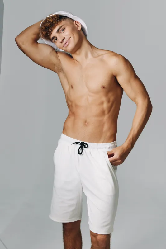 Young man with muscular body posing shirtless in white shorts against gray background. Fitness, confidence and casual style with hand behind head and relaxed expression in studio.