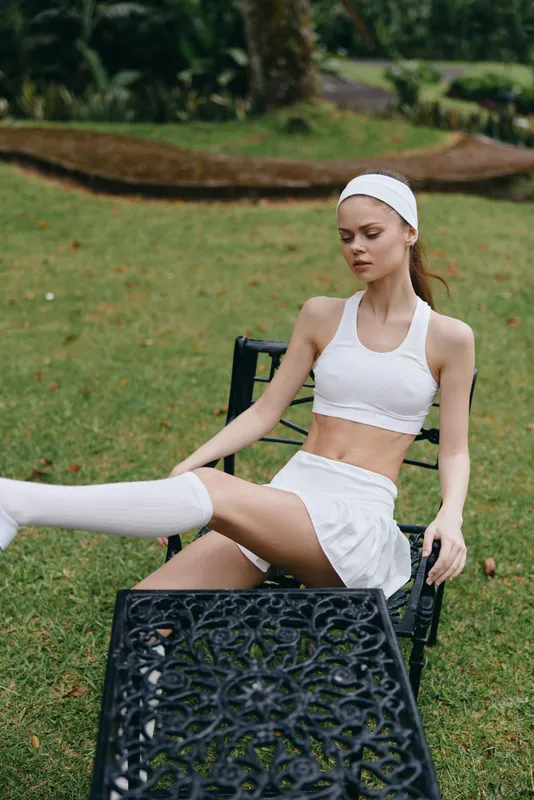 A fitness model in a white sporty outfit poses confidently on a garden chair, showcasing active lifestyle and healthy living in a vibrant outdoor setting