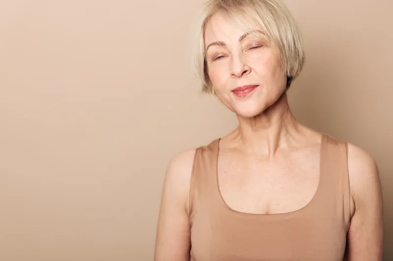 Blonde mature woman with short hair closes eyes and smiles gently. She wears a beige tank top against a neutral background. Concept of calm, confidence, natural aging and beauty.