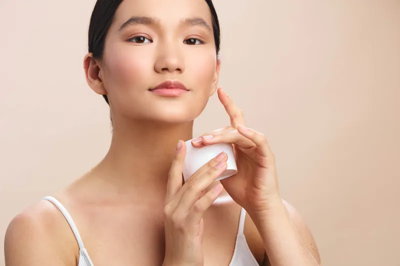 asian woman applies skincare product in a clean studio setting, presenting a calm, confident expression that conveys fresh beauty, self care, and a relaxed skincare routine.