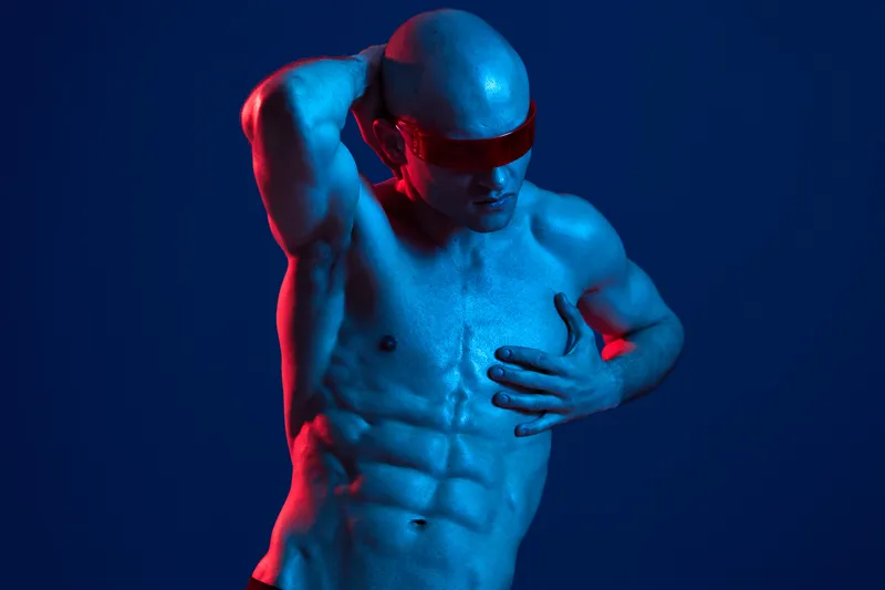 Athletic man poses dramatically against a blue backdrop, showcasing his toned physique and wearing stylish red accessories. This image emphasizes strength and creativity.