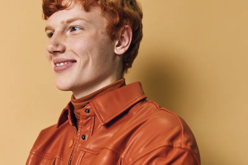 Young man with red hair wearing brown leather jacket looking sideways against plain background. Casual fashion portrait with confident smiling expression and natural light.