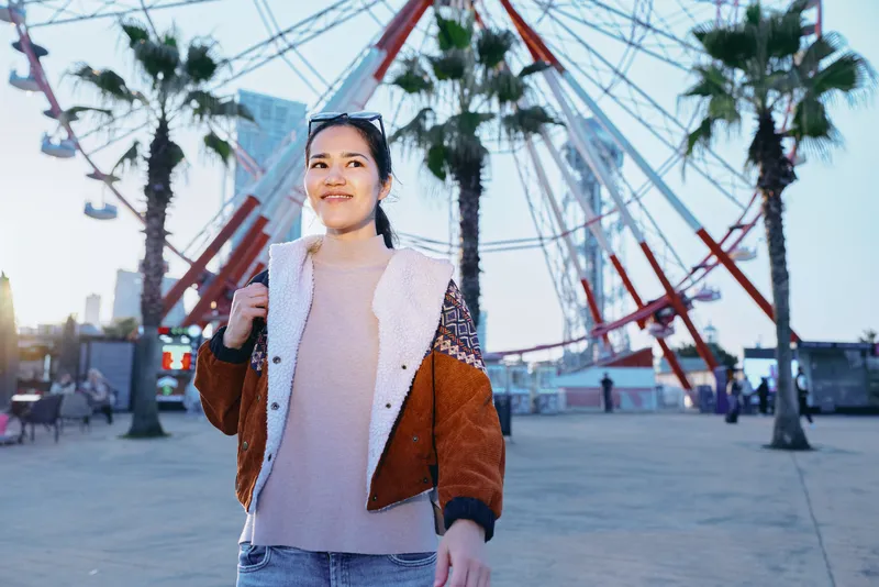 Smiling woman in a warm brown jacket and light sweater strolling along a coastal boardwalk with palm trees and a ferris wheel in the background, casual street style, urban leisure, candid fashion