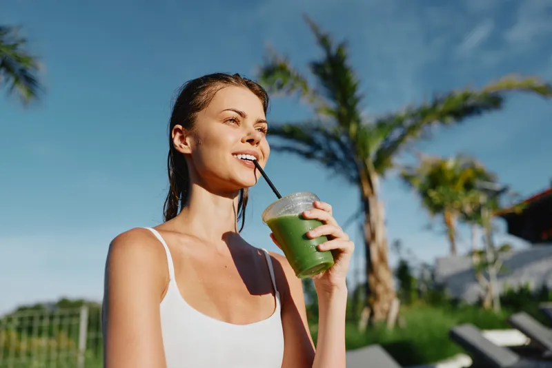 Candid lifestyle shot of a smiling woman enjoying a green smoothie outdoors on a sunny day with palm trees, casual white top, relaxed mood, and refreshing healthy beverage