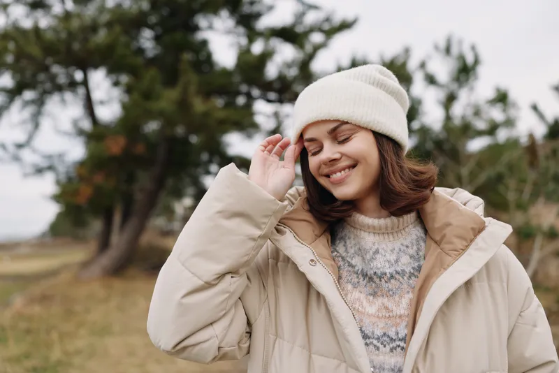 Woman in winter coat and beanie enjoying outdoor nature, smiling softly in a chilly field with trees in the background, cozy knit sweater and relaxed mood.