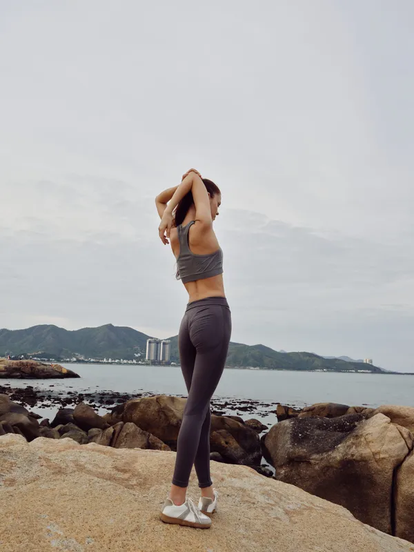 Athletic woman stretching on rocky shore, wearing grey sportswear, capturing a serene moment by the sea with mountains in the background, emphasizing fitness and wellness