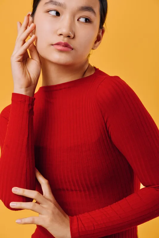 Young woman in red sweater posing with thoughtful expression on yellow background. Female model looking away with hand near face, casual fashion portrait with soft lighting and minimal style.