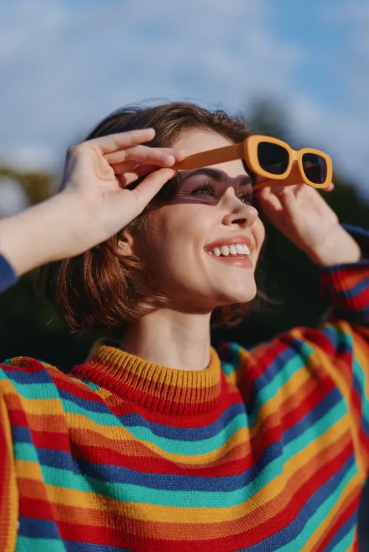 Woman holding yellow sunglasses and smiling in a colorful sweater, outdoor portrait of a happy fashion model with short hair enjoying sunny day and vibrant striped knitwear.