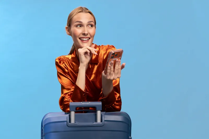 Happy young woman in shiny orange blouse stands behind blue suitcase holding smartphone, smiling and looking aside on blue background. Concept of travel, vacation, and modern technology usage.