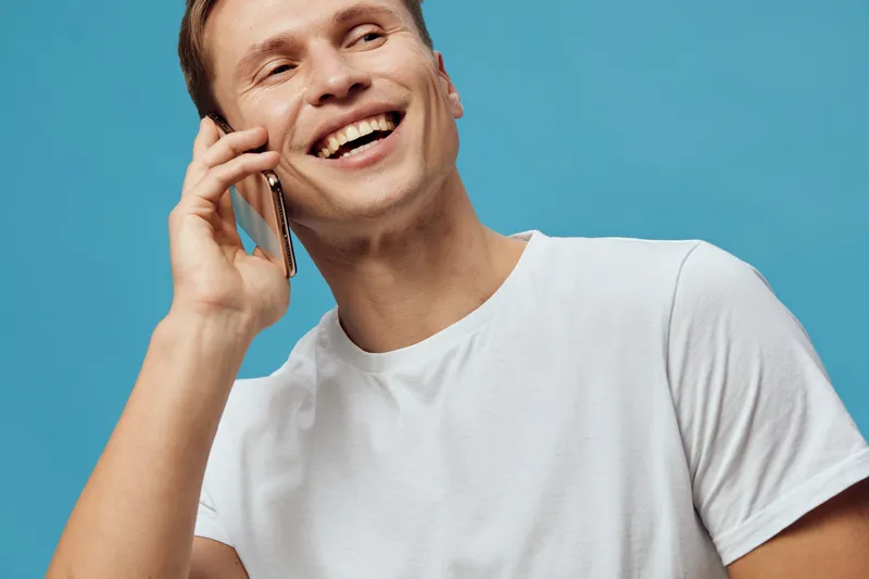 Smiling man cheerful young adult talking on mobile phone, happy expression, casual white t-shirt, blue background, male communication lifestyle image in studio shot. People concept