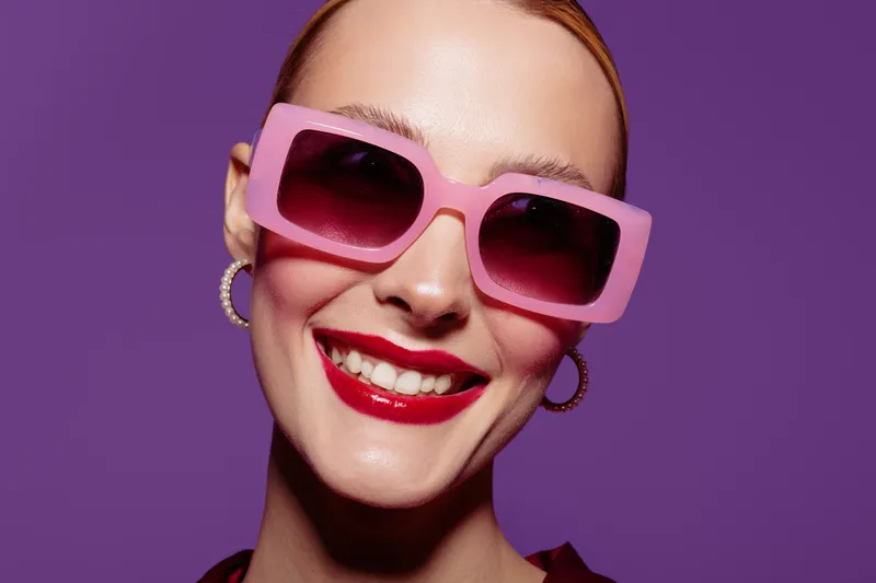 Close-up of a smiling woman wearing large pink sunglasses and red lipstick against a purple background. Concept of fashion, beauty, summer style, and confident mood.