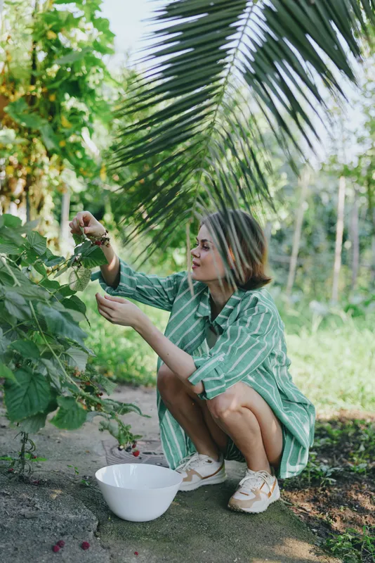 A woman harvests fruit in a sunny garden, crouching beside a bowl under lush foliage.