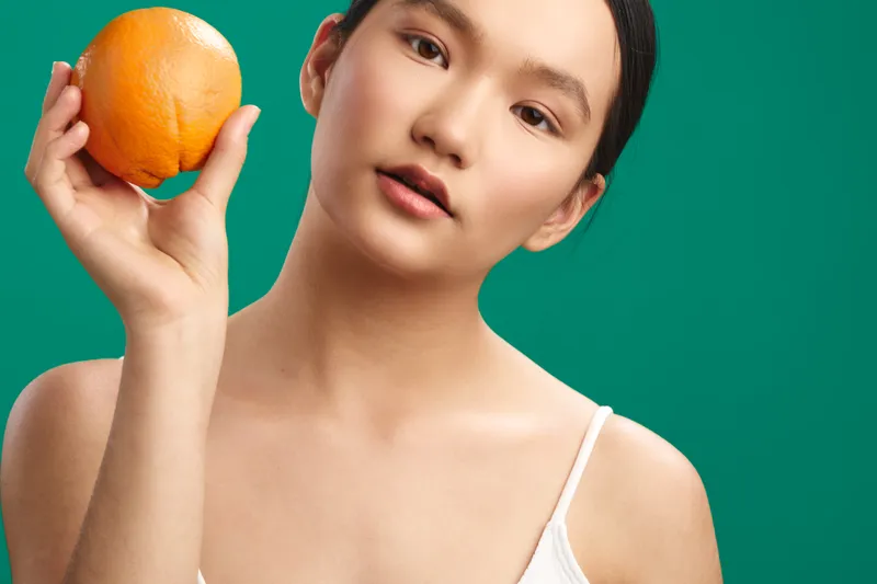 Asian woman holds an orange near her face against a vivid green backdrop, creating a fresh, minimalist portrait with soft lighting and a calm, confident expression.
