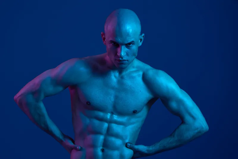 Strong man posing in blue lighting, showcasing his muscular physique with an intense expression, embodying strength and determination in a vibrant atmosphere.