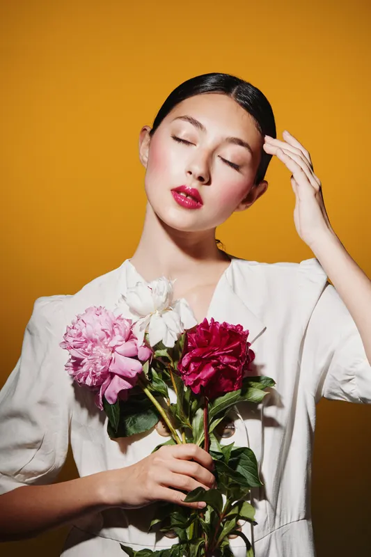 Beautiful woman with vibrant red lips and closed eyes holds colorful flowers. Studio portrait on bright orange background. Concept of beauty, fashion, and creativity.