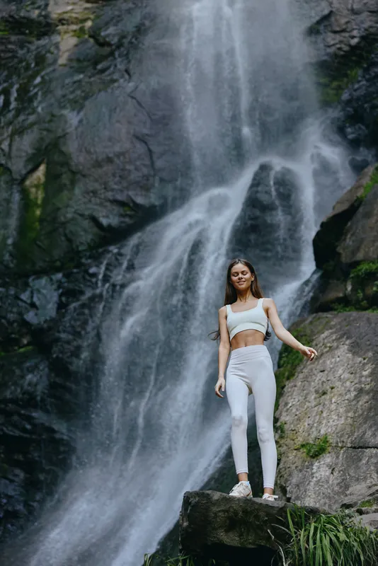 young woman standing near waterfall wearing white sportswear, natural outdoor setting, active lifestyle, serene and refreshing background