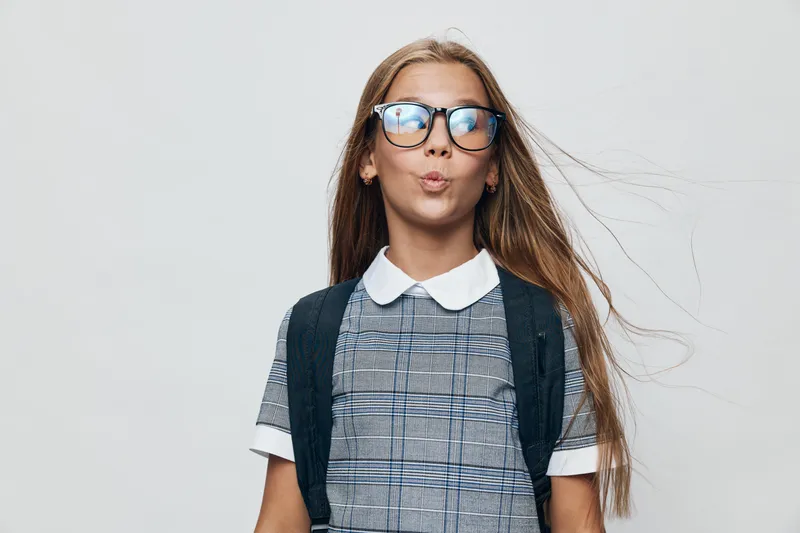 Girl with glasses and long hair wearing checkered dress and black backpack posing on white background. Youthful student in casual outfit with confident expression in studio setting.