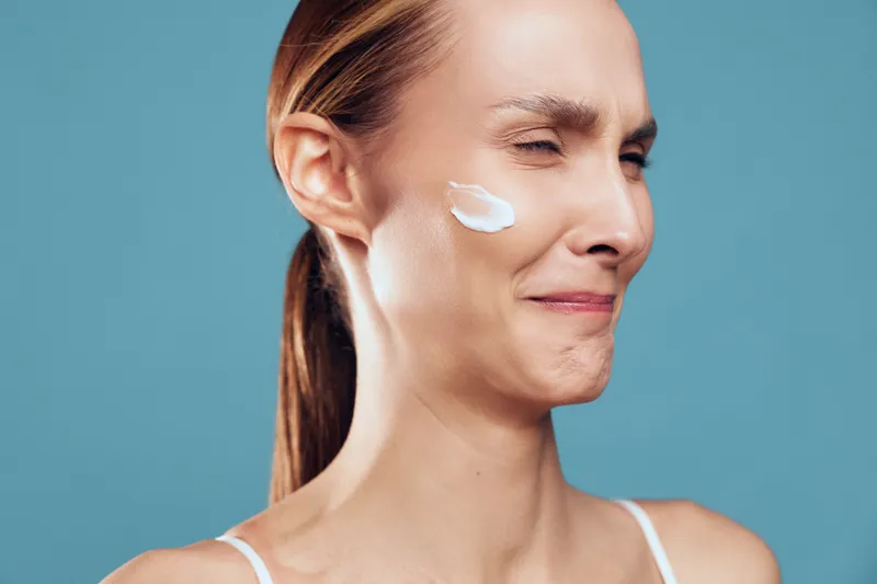 Young woman with a cream patch on her cheek smiles softly against a blue background, showcasing a simple skincare routine in a clean studio setting for promoting wellness and self care.