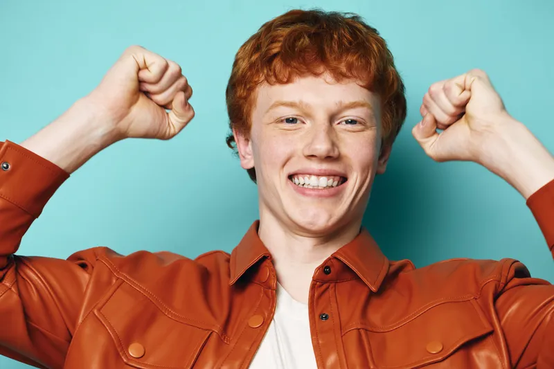 Young man with red hair and freckles smiling broadly and raising fists in celebration against turquoise background. Casual style and positive emotion of success and happiness.