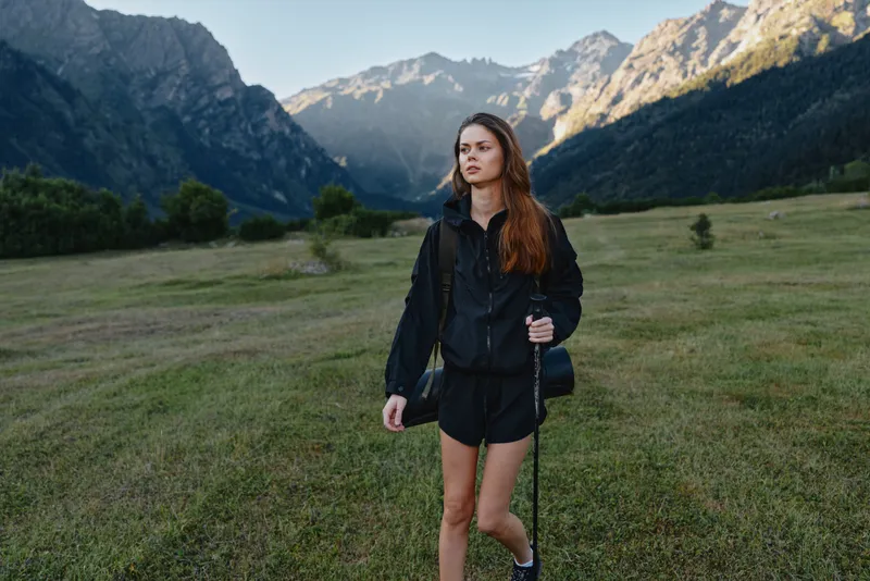 A young woman hiker stands in a wide meadow with distant mountains. She wears a black jacket and shorts, holds a trekking pole, and looks ahead with calm, adventurous focus.