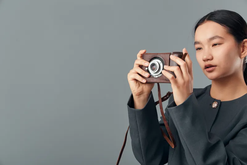 Woman holding vintage camera and composing photo with focused expression on gray background. Asian female photographer using retro style camera for creative shot indoors.