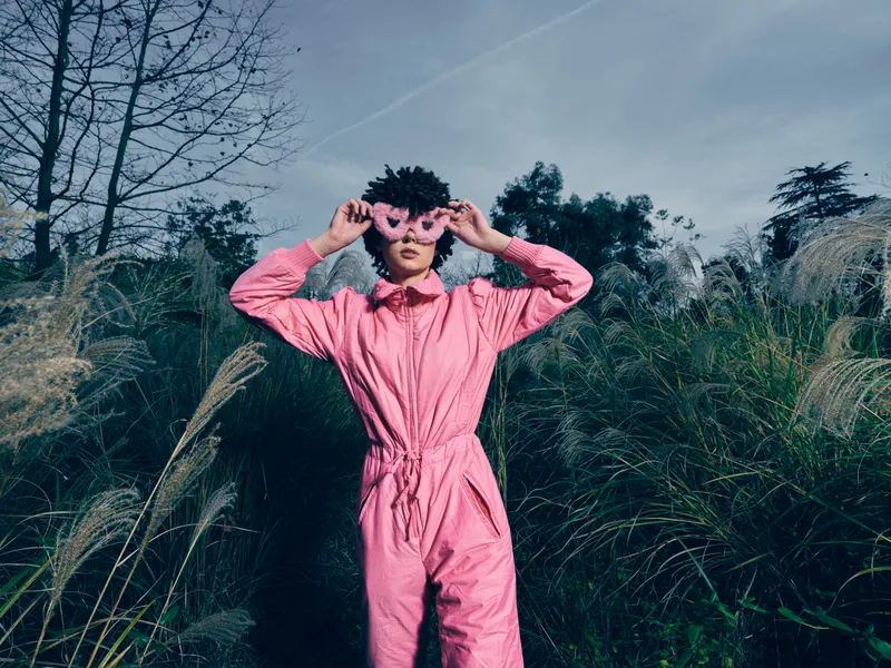 Outdoor portrait of a person in a pink jumpsuit, playful with round glasses, framing eyes among tall grasses and trees in natural light, bold fashion moment.