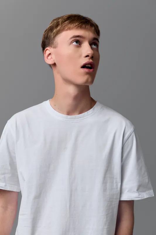 Man with surprised expression looking up, casual white tee, studio lighting, youth in simple attire, emotion and curiosity for fashion, lifestyle and stock usage