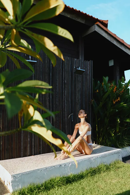 Young woman in white swimwear sitting on a sunlit terrace, enjoying warm sunlight, relaxed pose, tropical plants around, peaceful outdoor lifestyle moment.