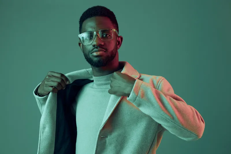 Stylish young Black man in glasses poses confidently, wearing a light grey coat against a vibrant green background, showcasing modern fashion and elegance