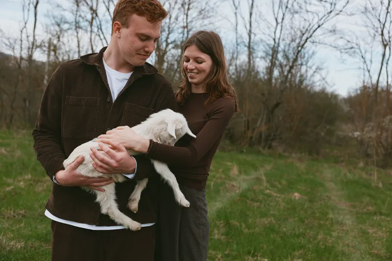 A young man gently holds a white baby goat while a young woman smiles standing beside him in a grassy outdoor area with bare trees in early spring.