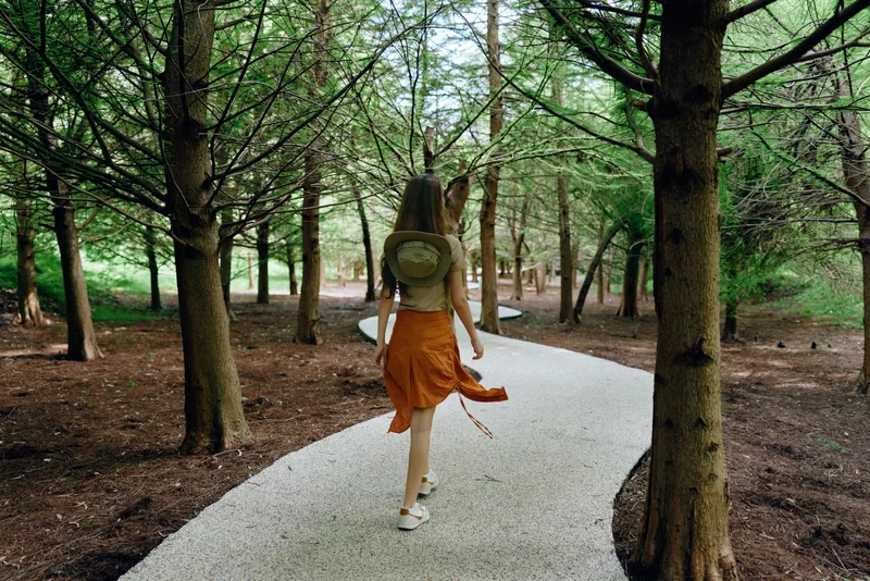 Woman walking on a winding path through a dense forest with a backpack and orange skirt, surrounded by tall trees and green nature on a peaceful woodland trail.