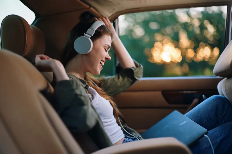 Young woman enjoying music in a car, wearing headphones, with a joyful expression, amidst a golden sunset background.