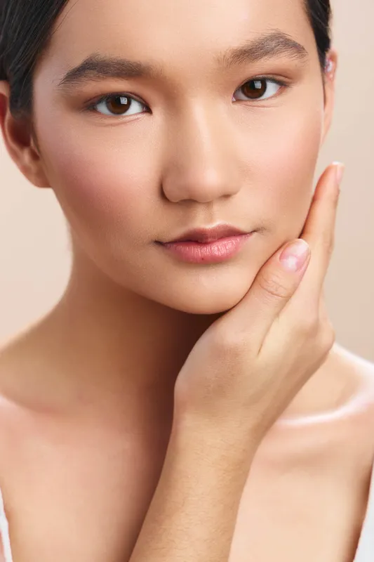 Asian woman poses with a calm expression, touching her face in a studio portrait. Soft lighting highlights smooth skin, creating an elegant and intimate beauty shot.
