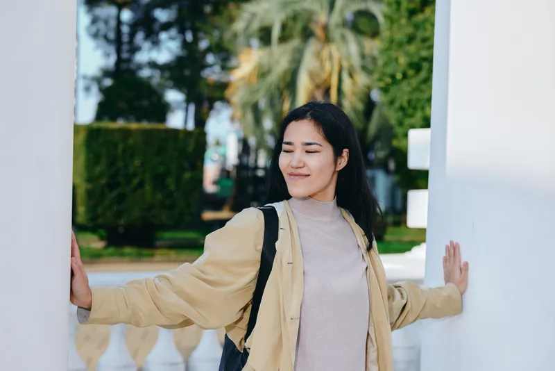 Woman with backpack stands between white columns outdoors, smiling softly in a beige jacket and light sweater, showcasing casual travel style in a sunlit park with trees nearby