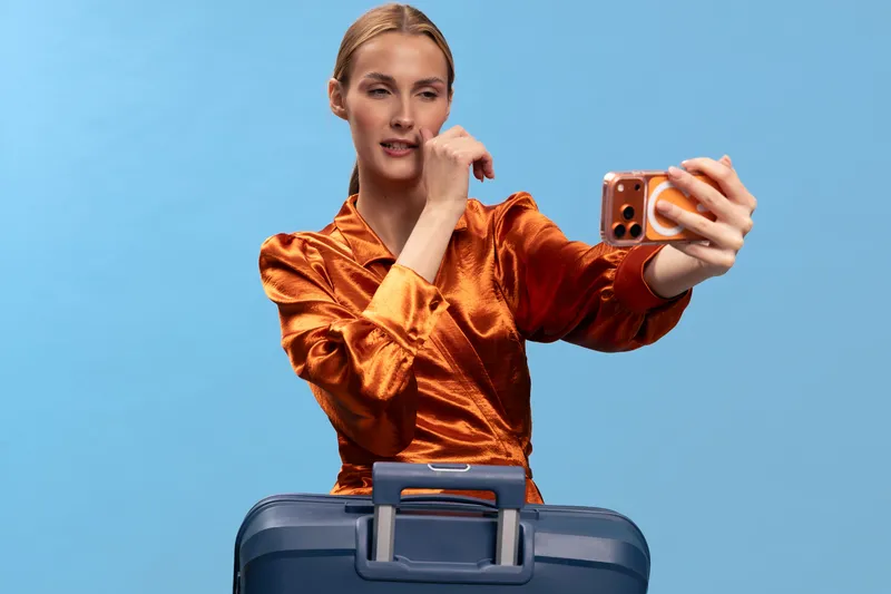 Young woman in orange satin blouse takes selfie with smartphone near blue suitcase on blue background. Concept for travel, vacation, lifestyle, and social media content creation.