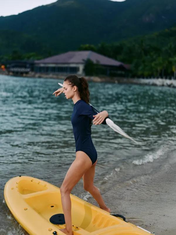 Young woman in a navy swimsuit steps onto a yellow kayak from a sandy shore, framed by calm water and distant mountains, her posture poised for balance and motion.
