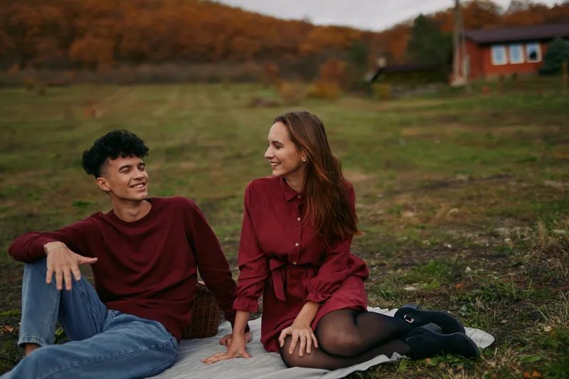 A relaxed young couple shares a candid outdoor moment on a blanket in an autumn meadow, conveying genuine connection and subtle authenticity through natural expressions.