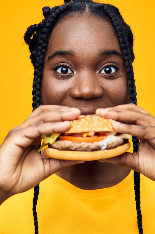 Joyful girl with braids holding a burger against a bright yellow background, celebrating delicious fast food in a vibrant setting