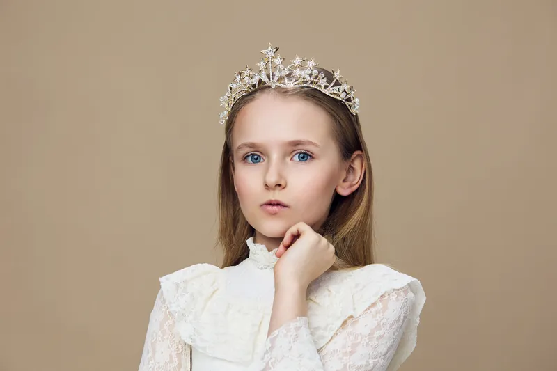 A young girl in a beautiful crown, wearing a delicate white dress, displaying an elegant expression against a soft beige background, perfect for portraits