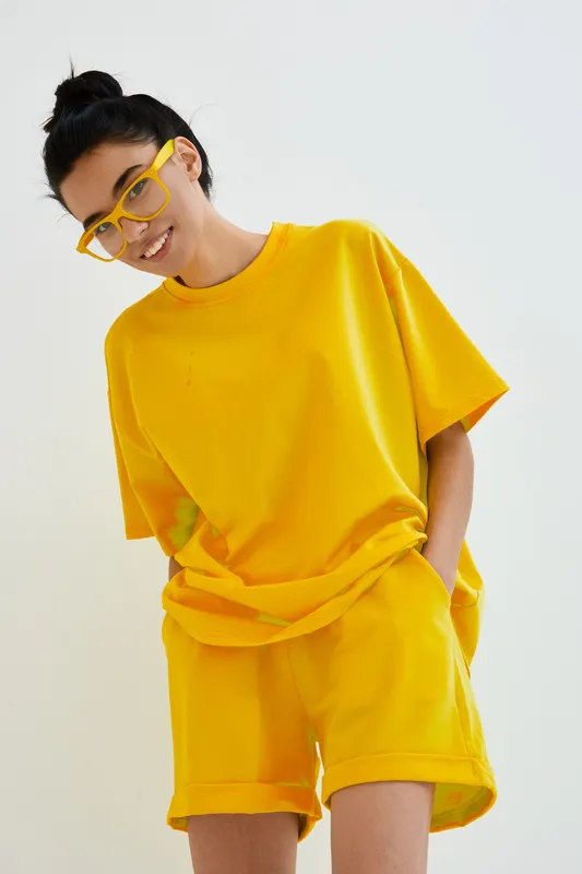 Young woman wearing yellow oversized t-shirt and shorts with matching yellow glasses smiling against bright white background in casual summer outfit.