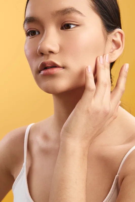 Asian woman portrait closeup with contemplative expression as she touches her face against a warm backdrop, conveying calm beauty and a soft, serene skincare focus in a gentle studio setting.