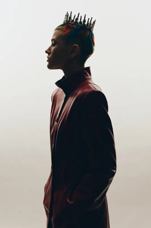 Profile of a woman with colorful hair and a crown wearing a dark jacket, standing against a neutral light background in a stylish and bold pose.