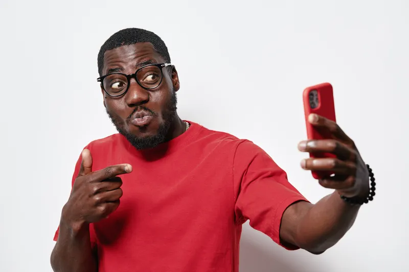 Black man with dark skin and glasses wearing red shirt taking selfie with smartphone making funny face on white background. African ethnicity, casual style and modern technology.