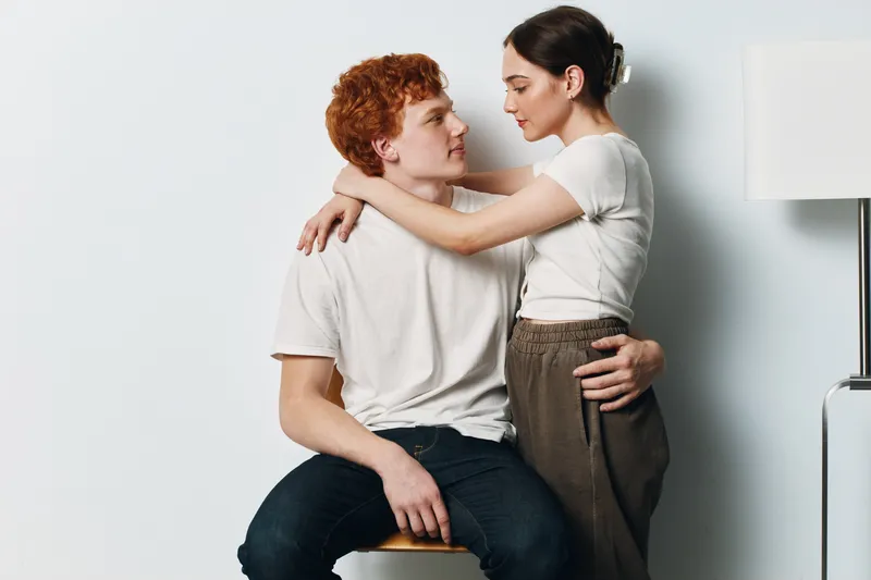 Couple embracing and looking at each other with affection in simple casual clothes. Young man sitting on a chair and woman standing with arms around him indoors.