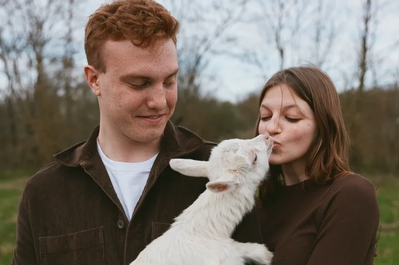 Couple outdoors share a tender moment with a playful goat kid, smiling softly as the animal nuzzles their faces. Natural light, rustic setting, wholesome lifestyle.